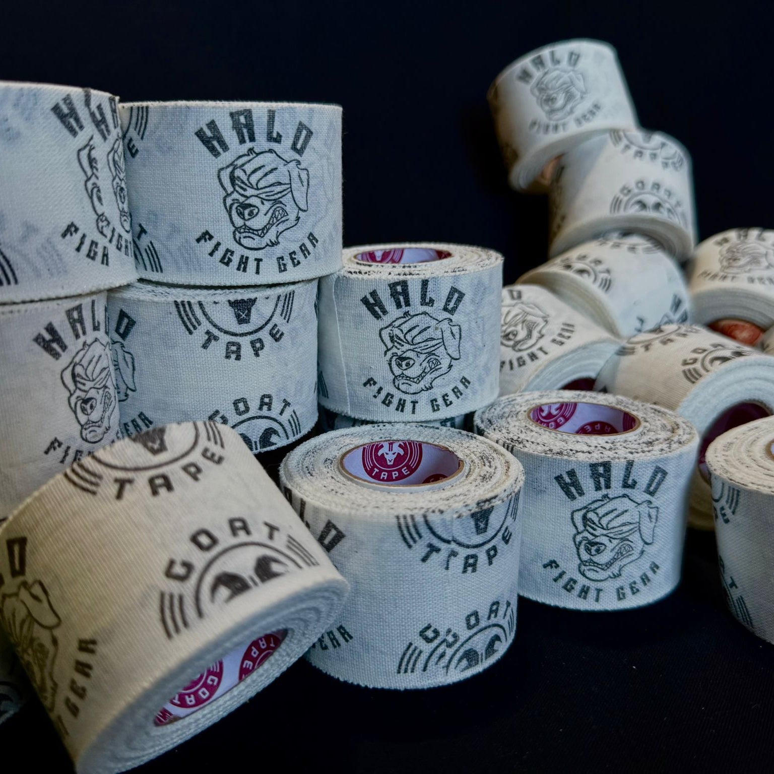 The image displays multiple rolls of athletic tape on a black surface. The white tape is printed with "HALO FIGHT GEAR" and "GOAT TAPE," accompanied by the logos for both brands. The inner cores have a red and white pattern. The rolls are arranged in stacks and side by side.